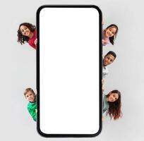 Diverse Kids Posing Near Huge Phone Blank Screen Hiding And Smiling To Camera Advertising Mobile Application Over Gray Studio Background. Children And Gadgets Concept. Square Shot, Mockup photo