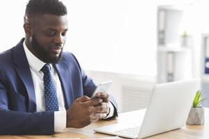 Handsome african businessman using cellphone at workplace, sending message or email, empty space photo