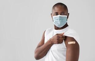 Covid-19 vaccine. Black man in protective mask showing an adhesive bandage on shoulder after injection. Patient standing over light studio background and looking at camera, copy space photo