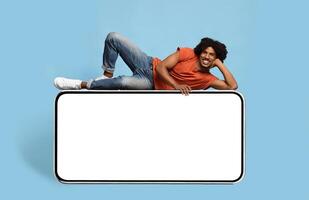Cheerful african american young man relaxing on huge horizontal brand new smartphone with empty screen and smiling at camera, mockup, black guy enjoying newest entertaining mobile app photo