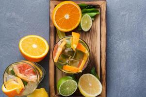 Summer citrus drinks background. Healthy detox water in glasses and fruits variety on wooden board on gray table. Colorful backdrop with oranges, lemon and lime, top view, copy space photo