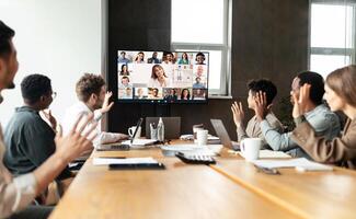 Distant Virtual Meeting Concept. Diverse business people making online call with colleagues, sitting at desk in boardroom, waving hands at tv screen at office, talking on web with remote workers photo