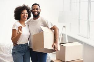 Real Estate And Family Housing. African American Couple Posing With Moving Boxes And New House Key Hugging Smiling To Camera Standing Indoors. Empty Space For Text photo