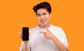 I Recommend This Website. Smiling young asian man holding smartphone with black empty screen in hand and pointing at device. Gadget with blank space for mockup, isolated over orange studio background photo