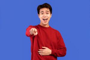 Joke And Prank Concept. Young asian guy mocking and laughing at you, standing isolated over blue studio background. Man pointing finger to the camera and touching belly, shame on you photo