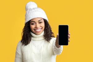Modern Technology Concept. Smiling african american woman in winter hat holding cellphone with empty black screen over orange studio background. Happy lady showing gadget display for mock up photo