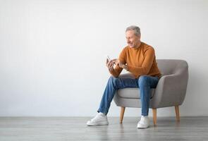 Cheerful Mature Guy Using Mobile Phone Testing New Application Sitting In Armchair Isolated On White Studio Wall Background. Gadgets And Mobile Communication Concept. Free Empty Copy Space For Text photo
