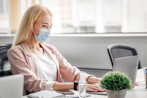 New normal, modern office protected from virus and managers job. Blonde business woman in protective mask typing on computer on table with bottle of antiseptic, working after quarantine, copy space photo