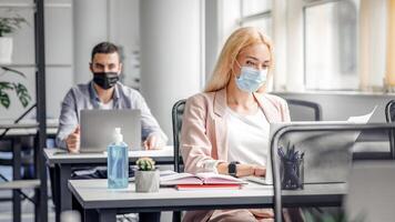 New working conditions and social distancing after quarantine. Blonde woman in protective mask works with documents and laptop at workplace with notepad and big bottle of antiseptic in interior photo