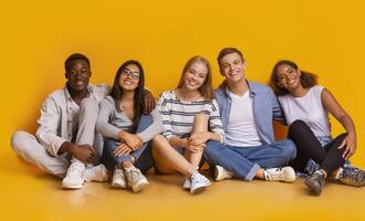 Multiracial joyful students sitting on floor, embracing and having fun over yellow studio background, students lifestyle concept, panorama photo
