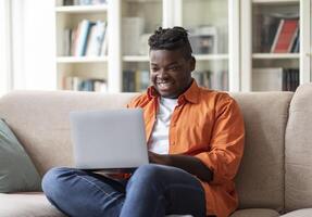 Freelance Career. Happy Overweight African American Guy Using Laptop Computer Sitting On Couch At Home, Browsing Internet And Working Distantly Online. Remote Job, Website, Copy Space photo
