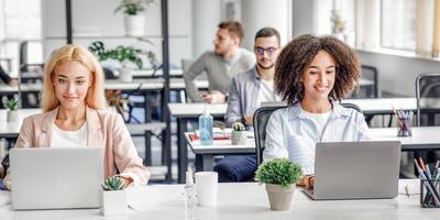 Work in modern office, digital technology and social distancing after quarantine. European and african american smiling women working at laptops, sits at table with antiseptics in interior, empty space photo
