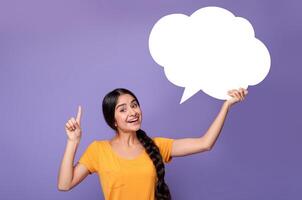 Inspired young indian lady having idea, holding blank speech bubble and pointing finger up, showing free place for your text, standing over purple background in studio, copy empty space photo