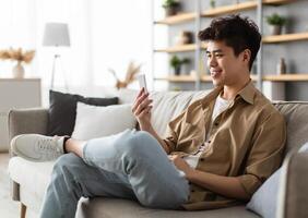 Cool Gadget And Application. Portrait of young smiling asian man holding mobile phone, typing sms message, sitting on the sofa in living room. Guy browsing internet, surfing web, using app, free space photo