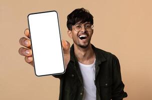 Emotional handsome hindu guy wearing eyeglasses showing brand new cell phone with white empty screen and smiling, recommending nice online offer, mockup, beige studio background photo