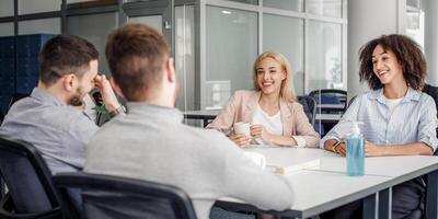 Coffee break with colleagues in rest room during quarantine covid-19. Smiling multiracial colleagues with cups talking through glass board and sitting at table with antiseptic in interior, copy space photo