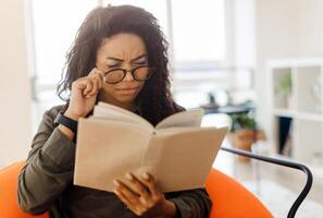 Focused confused young black lady trying to read paper book, squinting to see more clearly, wearing glasses, having difficulties seeing text because of vision problems, sitting on pouf in living room photo