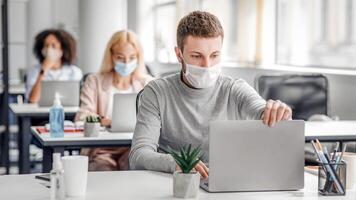 Coronavirus quarantine and office work with colleagues keeping social distancing. Portrait of young man in protective mask make call with client at table with laptops with antiseptic photo