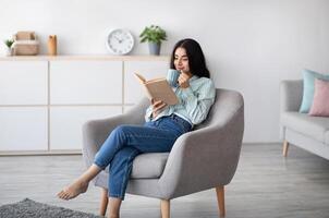 Attractive Indian lady with cup of aromatic coffee and open book relaxing in armchair at home. Pretty millennial woman enjoying hot beverage and reading exciting story, having lazy weekend indoors photo