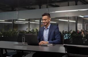 Positive young arabic manager working on laptop at modern hi-tech office, sitting at table, typing on computer keyboard, sending emails, communicating with business partners, copy space photo