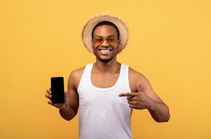 Black guy in summer outfit pointing at cellphone with blank screen on yellow studio background, space for mobile application or website. African American man showing smartphone with mockup for your ad photo