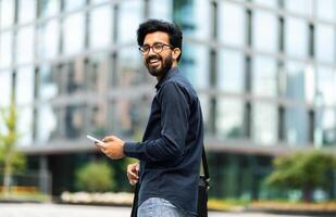 Happy handsome young middle eastern guy in casual and eyeglasses IT professional going to office, walk by street, using phone, checking email, using business mobile app, smiling at camera, copy space photo
