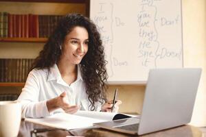 Online Conference. Portrait of professional female teacher in wireless earbuds giving virtual lecture, making call and talking with students, sitting at table near whiteboard with grammar rules photo