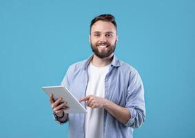 Modern technologies and communication. Portrait of happy bearded man using tablet computer over blue studio background. Young guy studying or working online on touch pad photo
