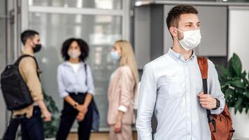 Social distance and work in office after covid-19 quarantine. Focus on millennial man with protective mask and backpack, looks to the side in hallway, and workers are blurred, free space photo