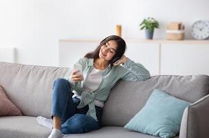 Joyful Indian lady in headphones sitting on couch and listening to music on smartphone at home, full length. Charming young woman enjoying her favorite song or audio book indoors photo