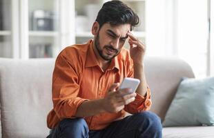 Stressed upset arab man in casual sitting on couch at home, holding cell phone, looking at gadget screen and touching his head, reading bad news, reading message, copy space photo