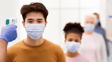 Temperature Screening. Hand With Contactless Themometer Measuring Fever, Scanning Forehead Of Chinese Man Standing In Queue Indoors. Coronavirus, Covid-19 Virus Spread Control. Panorama, Copy Space photo