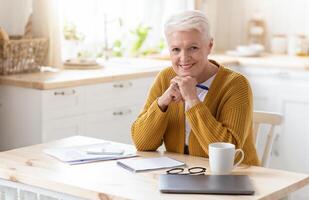 Positive elderly lady freelancer working from home or studying, sitting in kitchen with laptop and smartphone, taking notes while reading financial report, smiling at camera, copy space photo