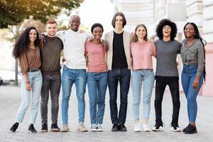 Friendship, community, team concept. Multiracial group of happy millennial men and women posing on the street, standing in a row, embracing and cheerfully smiling at camera, full length shot photo