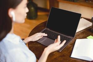 Online Call And Communication. Over the shoulder view of woman wearing earbuds, sitting at table, using laptop with black empty screen for virtual conference, talking to webcam, selective focus photo