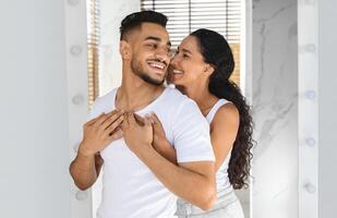 Portrait Of Happy Romantic Middle Eastern Couple Embracing While Standing Near Mirror In Bathroom, Cheerful Millennial Arab Spouses Hugging And Having Fun While Making Morning Beauty Routine Together photo