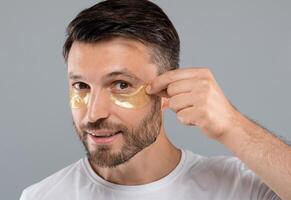 Excited attractive man applying eye patches on his face, closeup. Middle-aged bearded man with golden patches under eyes from wrinkles and dark circles, grey studio background, face care for men photo