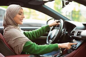 Car Playlist. Side View Portrait Of Muslim Woman In Hijab Driving Car And Listening To Music On Auto Audio System, Checking Automobile Options During Test Drive, Pushing Button On Dashboard Panel photo
