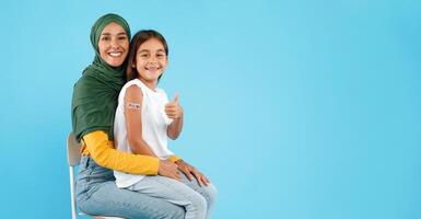 Covid-19 Vaccination For Children. Vaccinated Muslim Mother And Little Daughter With Plaster On Arm After Vaccine Injection Gesturing Thumbs Up Sitting On Blue Background. Panorama, Copy Space photo