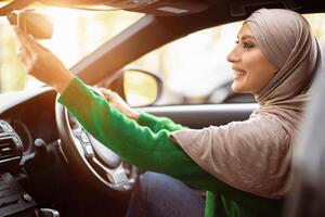 Buying New Car. Satisfied Muslim Lady In Hijab Adjusting Rear View Mirror, Testing Auto, Enjoying Her New Vehicle, Sitting On Driver's Seat In Modern Automobile, Side View With Sun Flare Light photo