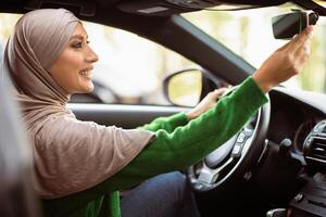 Side view portrait of smiling muslim woman in hijab adjusting rearview mirror while sitting in her car, selective focus. Happy Middle East lady in headscarf test driving new automobile photo
