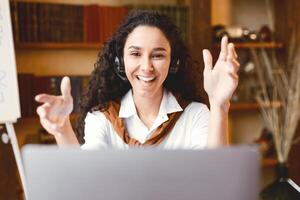 Teleconference And Telecommuting. Portrait of smiling woman sitting at desk, having call on laptop, explaining lesson to students or working and talking to colleagues and gesturing to webcam photo