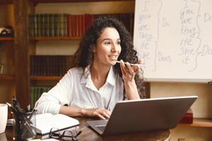 Voice Search And Communication Concept. Portrait of young curly lady sitting at table with computer, talking on smartphone, using laptop and virtual assistant to record message or making call photo