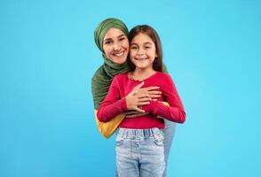 Cheerful Islamic Mother Wearing Hijab Hugging Her Little Daughter Smiling To Camera Posing On Blue Background. Arabic Mom And Her Kid Girl Standing And Embracing Together In Studio photo