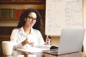 Remote Lecture Concept. Portrait of smiling young female tutor wearing glasses and wireless earbuds making online call using laptop, talking to webcam, asking students, whiteboard in background photo