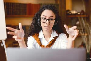 Remote Learning. Female English teacher having conference call with students and class group, using computer. Woman wearing eyeglasses and wireless headset, talking to webcam, explaining photo