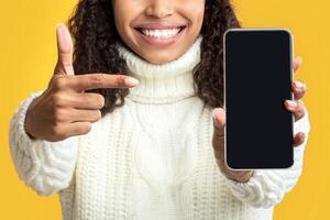 Mobile Application. Black lady in winter hat holding and pointing at empty cellphone screen over orange studio background. Happy woman showing and indicating device display for mock up photo