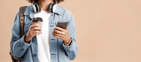 New message in online mobile application. Smiling african american female in headphones with smartwatch and cup of coffee holds phone and chatting with followers isolated on yellow background, cropped photo