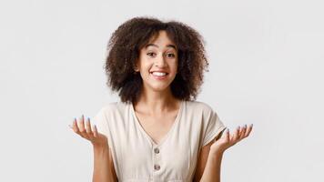 I do not know, can not choose or proposal. Millennial confused african american female student with curly hair in casual, smiles and spreads arms to sides, isolated on light background, free space photo