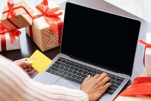 Ordering Presents. Over the shoulder view of african american female customer in protective face mask holding credit card and using computer with black empty screen with mockup, gift boxes on table photo
